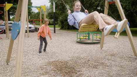 niña con síndrome de down con capucha balanceándose en un columpio en el parque en un día de viento. su amigo masculino empujándola