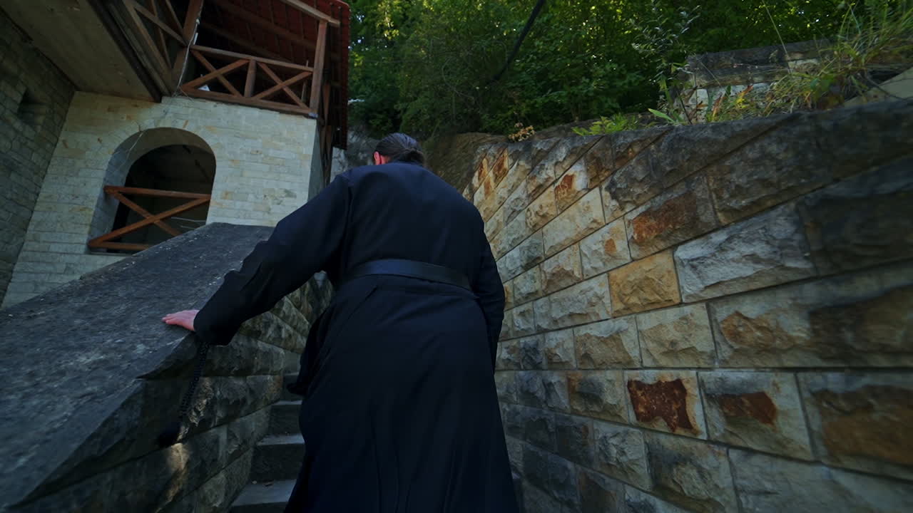 Rear view of a monk in black robe walking up the stone stairs. Priest in Orthodox monastery.