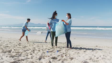 Diverse-group-of-women-wearing-volunteer-t-shirts-and-face-masks-picking-up-rubbish-from-beach