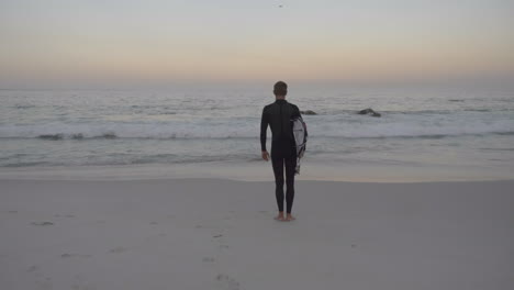 surfer standing on beach at sunrise holding surfboard in cape town south africa