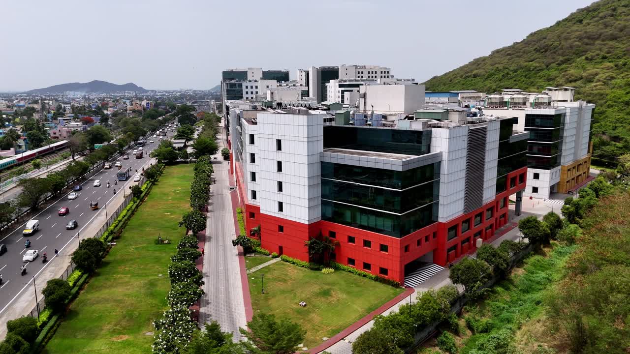 A modern, glass corporate building in Chennai is shown next to a busy highway and elevated railway. This scene captures the vibrant intersection of technology and infrastructure development