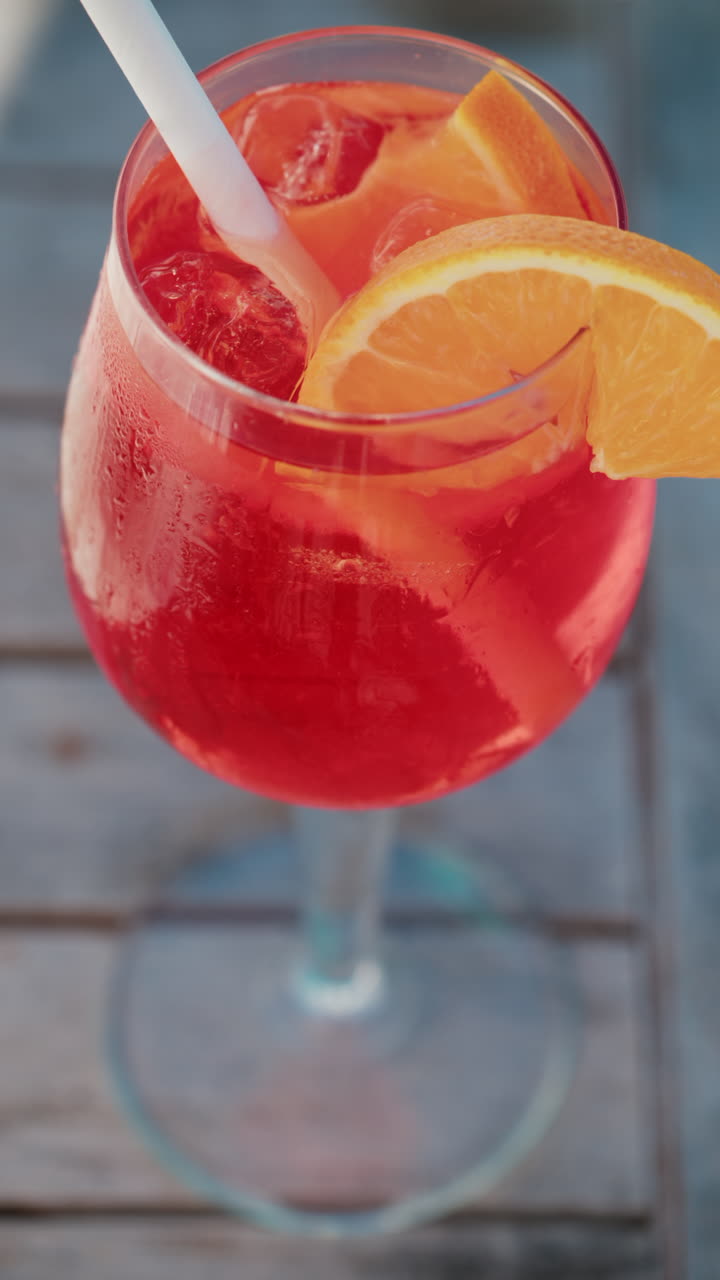 Primo piano di un cocktail rinfrescante rosso-arancio con fetta d'arancia e cubetti di ghiaccio, realizzato all'aperto in una giornata di sole a Cannes. Verticale
