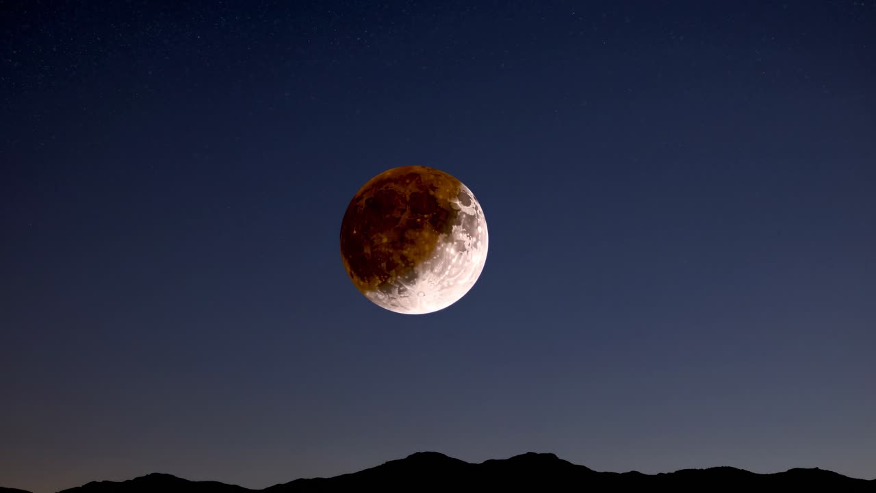 A wide-angle shot captures a lunar eclipse over silhouetted mountains under a starry sky