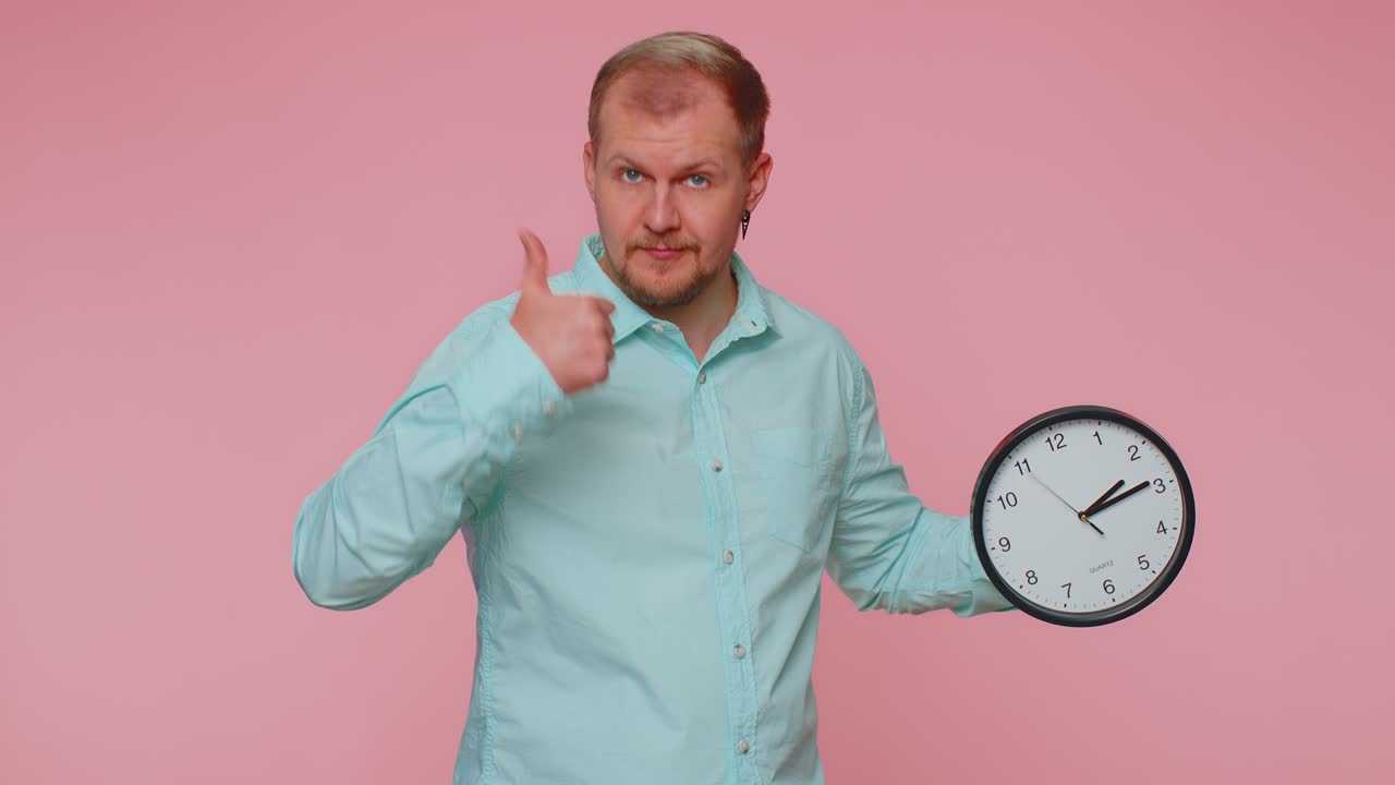 Bearded young man showing time on clock watch ok thumb up approve pointing finger at camera