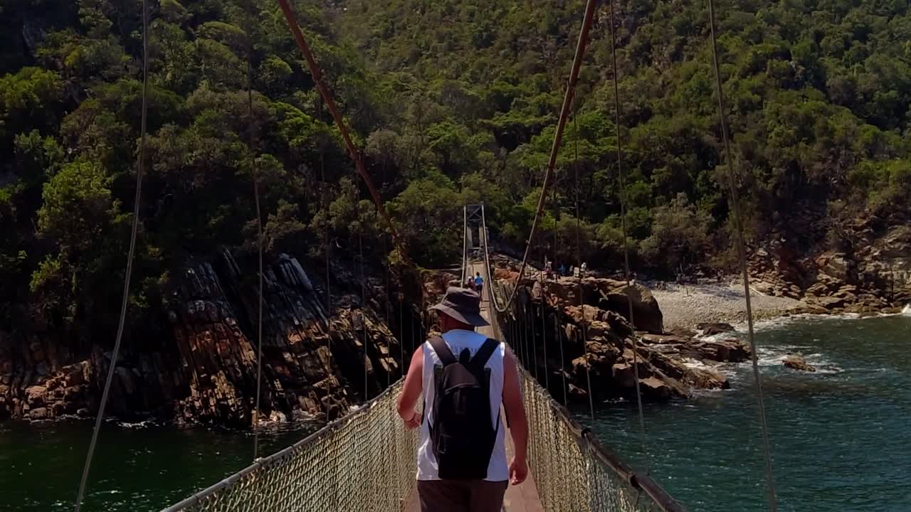 Um plano médio de um alpinista com uma mochila e chapéu de sol atravessando a famosa ponte suspensa na foz do rio Storms