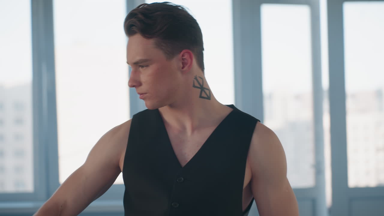 young man in black vest stylishly removes belt and hangs it around neck while performing fluid dance moves in sunlit studio with large window backlight