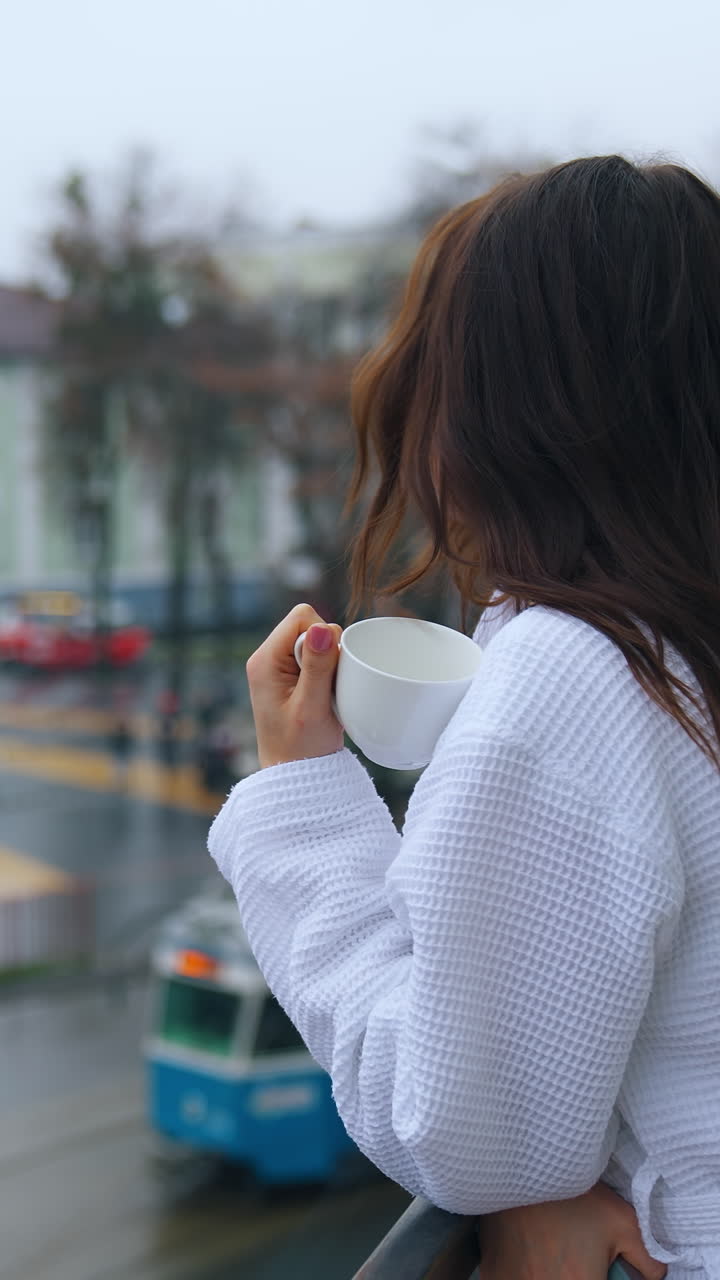 Beautiful girl drinking coffee on a balcony. Young woman in white dressing gown enjoys morning on a hotel balcony on city transport background. Vertical video