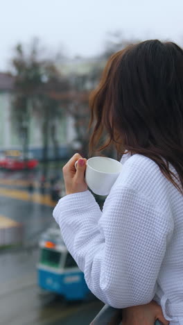 beautiful girl drinking coffee on a balcony. young woman in white dressing gown enjoys morning on a hotel balcony on city transport background. vertical video