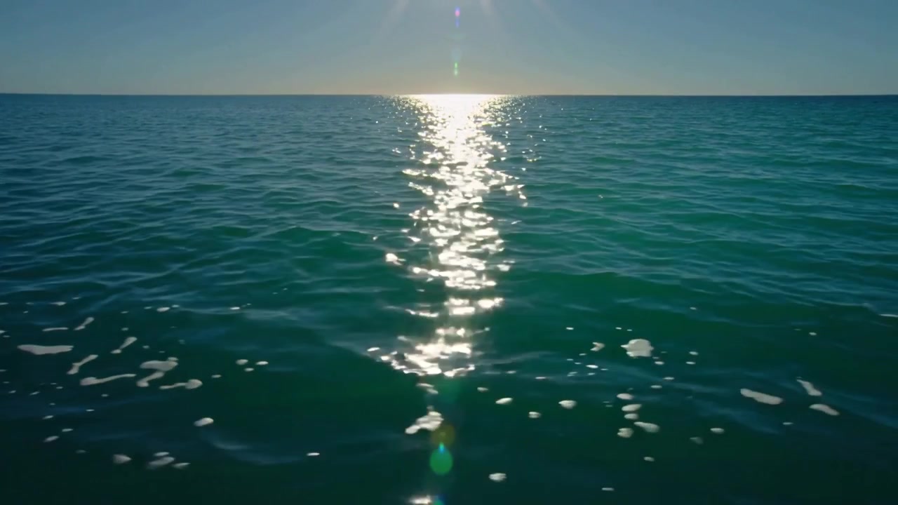 Drifting camera capturing low sun reflection on open ocean, revealing shifting foam and glint