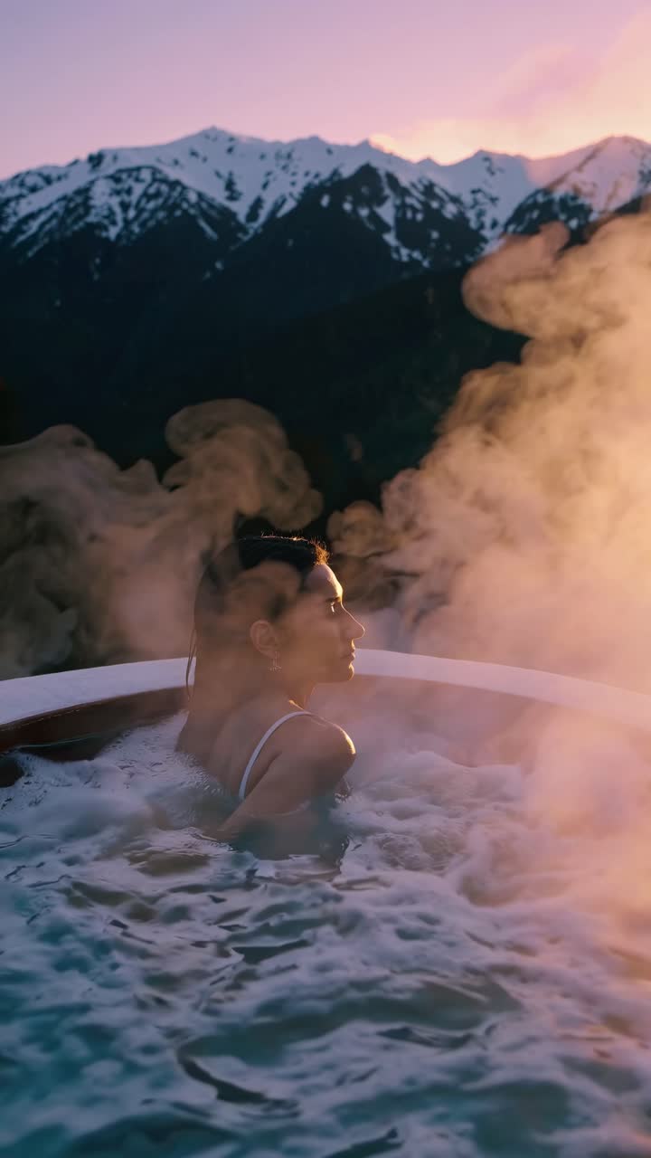 A serene video scene of a person in a hot tub with steam rising, set against snow-capped mountains
