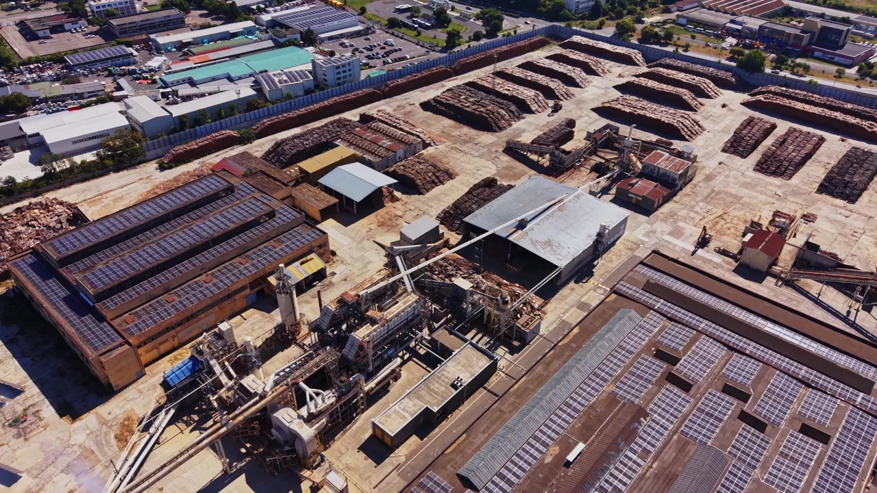 Un impianto di lavorazione del legno mostra grandi cataste e macchinari in un'area industriale.