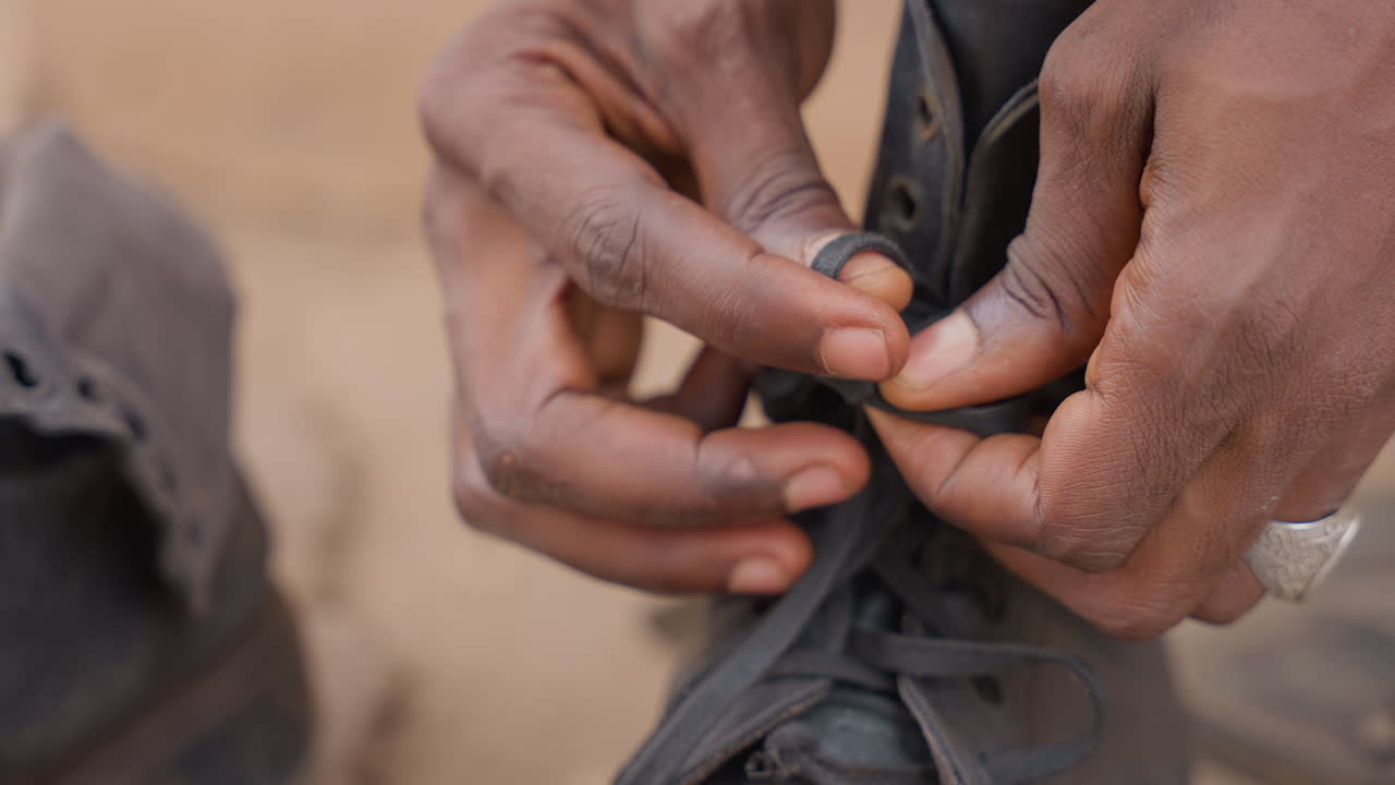 Black man threading shoelace with careful fingers outside house, closeup on hands and aged leather, aglet manipulation and patient knot forming, small maintenance task before daily chores, artisanal