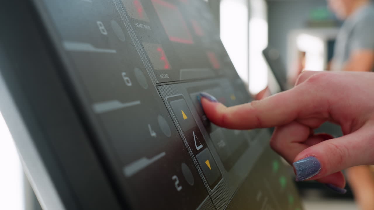 close up treadmill control panel with visible speed and incline indicators while user operates machine, blurred background reveals another person approaching treadmill under bright gym lighting