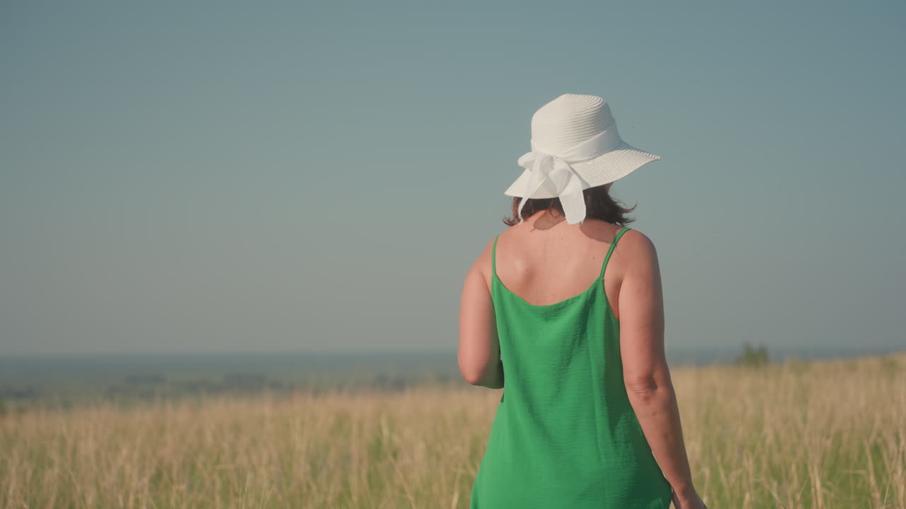 Elegant woman in white sunhat and green dress walking through tall dry grass under soft blue sky, back turned toward horizon, peaceful summer atmosphere captured in quiet countryside setting