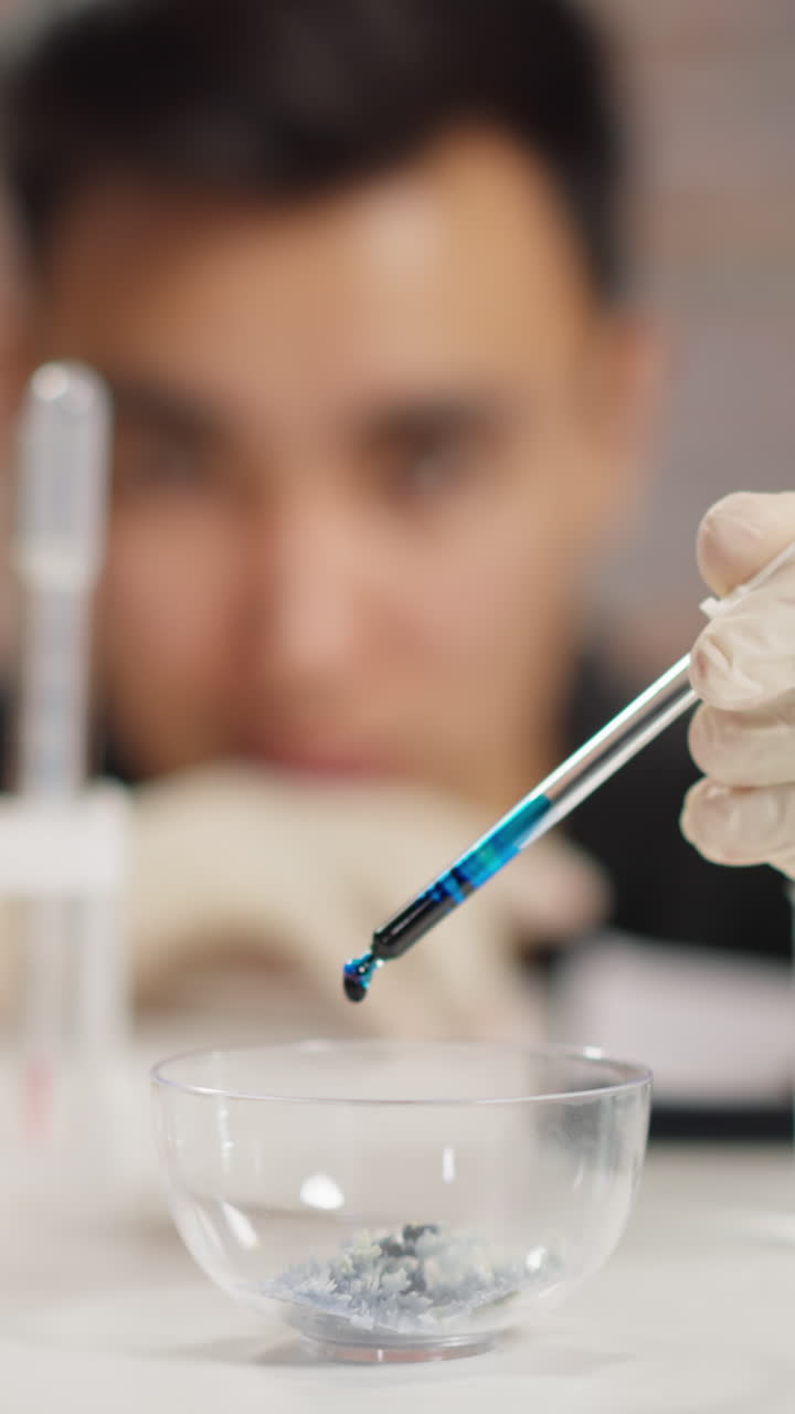 Man chemist with white gloves drops dark blue liquid reagent into bowl with crystalline substance at testing in scientific laboratory closeup