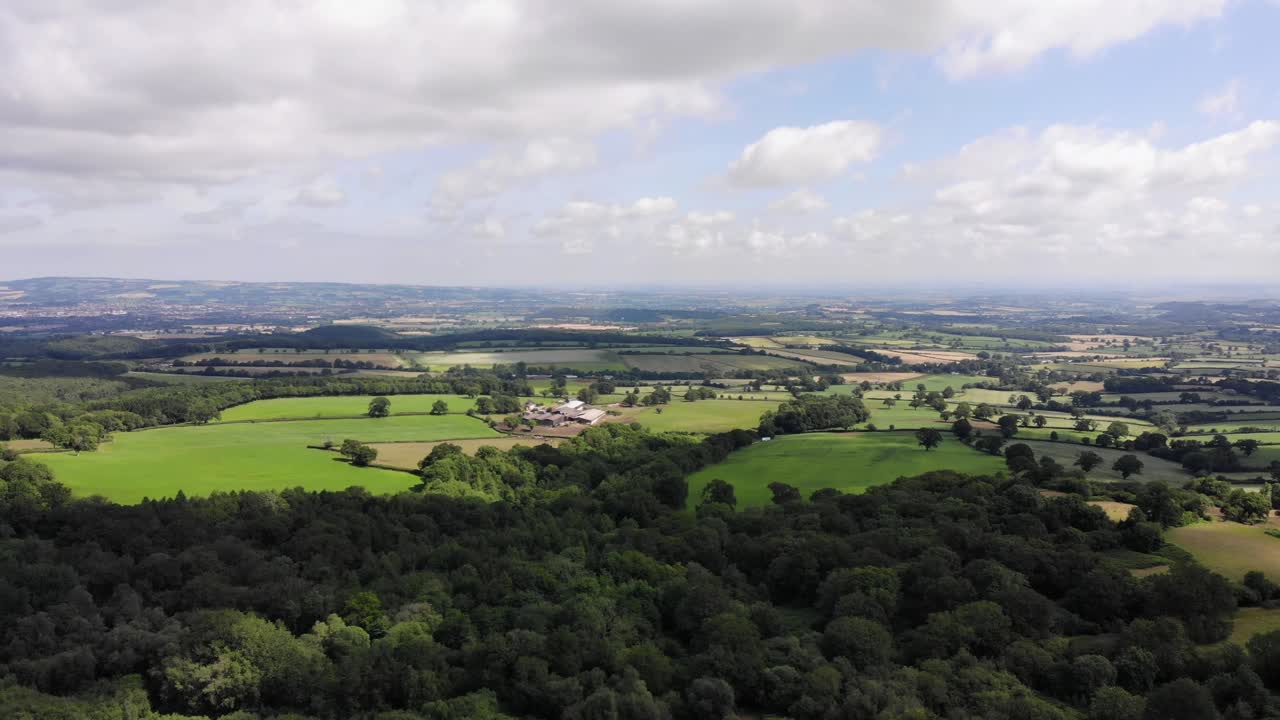 Aerial view of the Blackdown Hills countryside showcasing lush greenery, dense forests, and picturesque open fields under a partly cloudy sky. Panning Shot