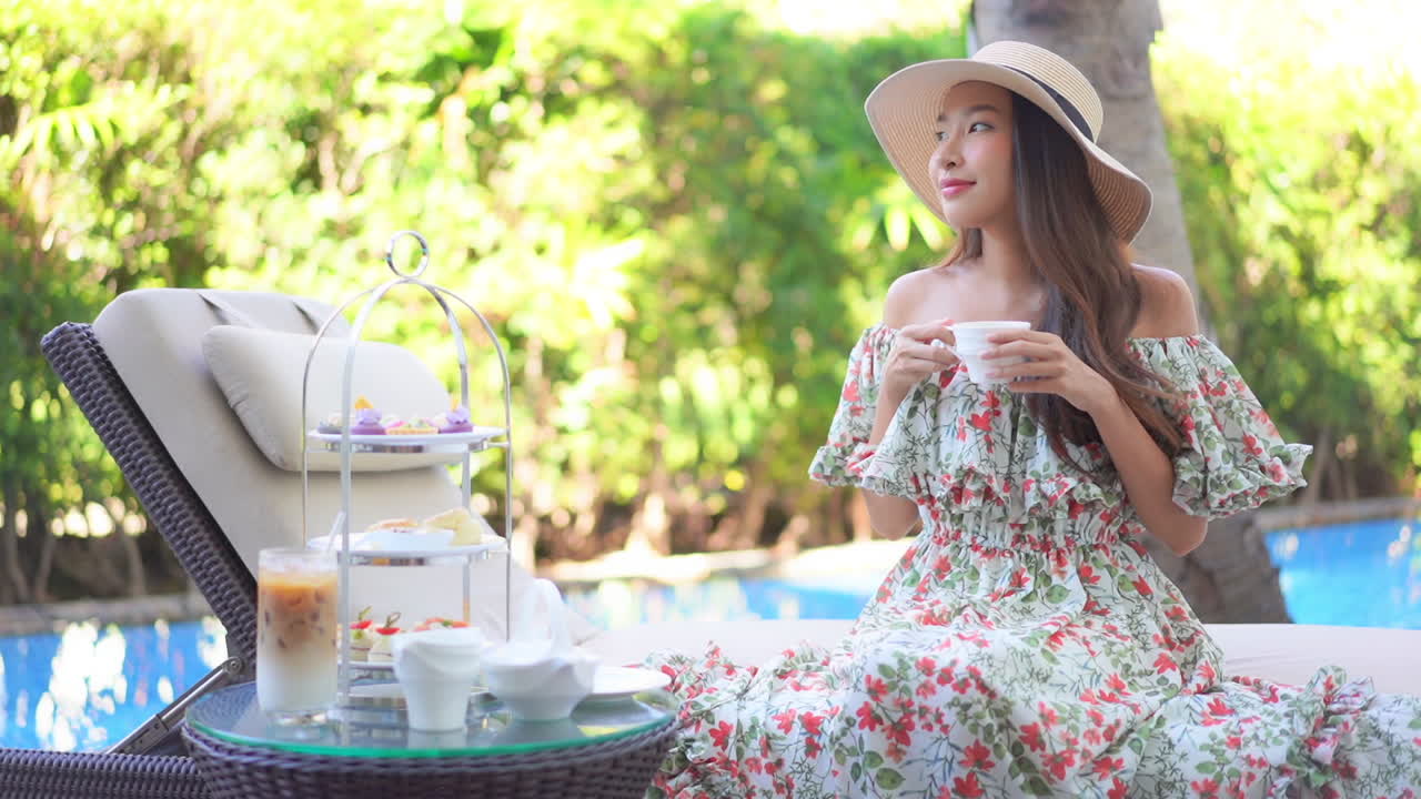 Beautiful Asian woman sits on deck-chair and drinking morning coffee near swimming pool resort