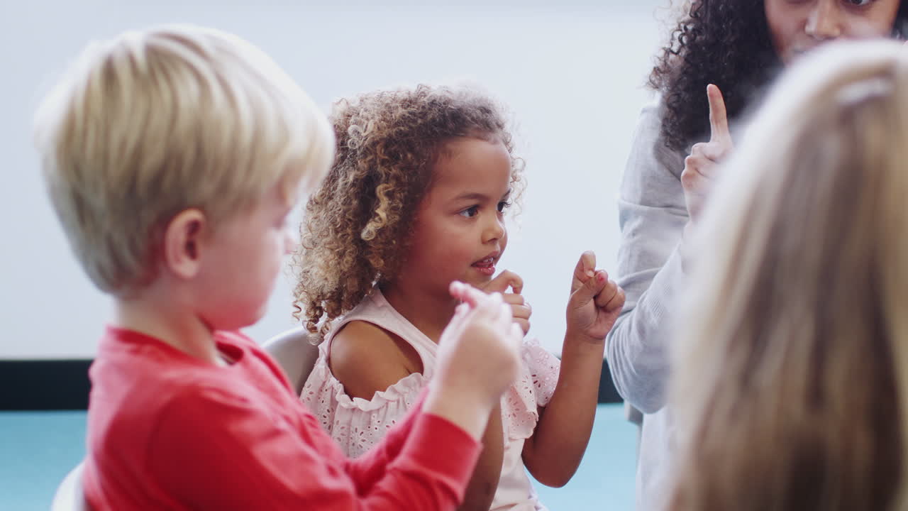 Bambini della scuola dell'infanzia in classe che alzano le mani, imparando a contare con la maestra, vista laterale.