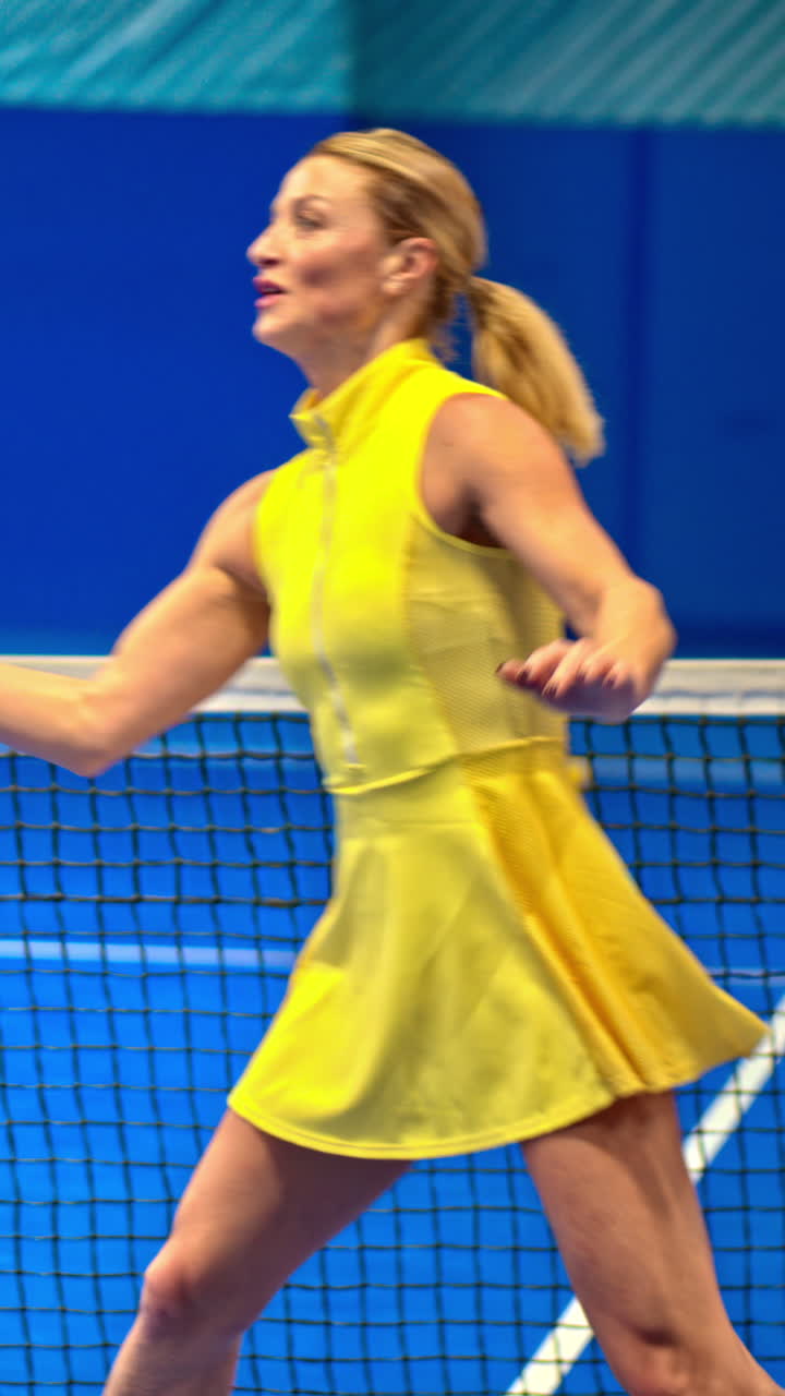 Woman in a yellow dress training to play pickleball on a blue, inside court. Vertical