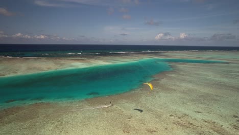 Un-Solo-Kitesurfista-Deslizándose-Sobre-Aguas-Cristalinas-De-Color-Turquesa-Cerca-De-Un-Cayo-Durante-El-Día,-Vibrante-Y-Serena-Vista-Aérea