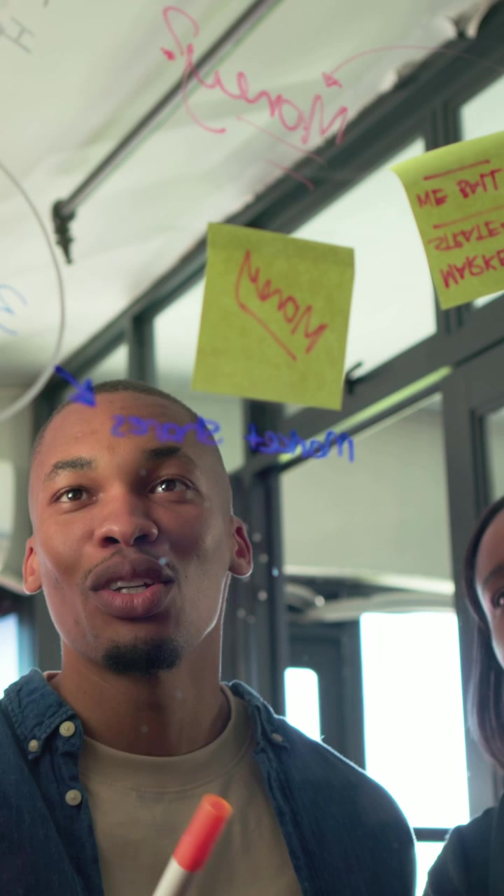 Vertical video: Pointing woman in beige blouse planning on glass at office, man adding sticky notes