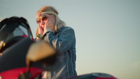 a woman wearing sunglasses and a flower headband is dancing with a joyful expression, moving her hands close to her face while standing near a red motorcycle in an open field