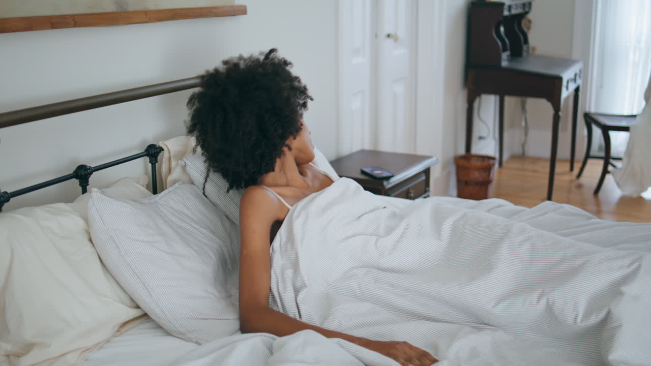 Ragazza assonnata si sveglia a letto. Donna africana prende il cellulare sdraiata sul materasso.