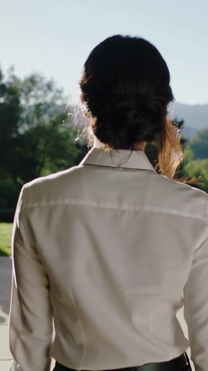 Vertical video: Stepping woman blouse low chignon via arch to lawn, sun drawing view of hills
