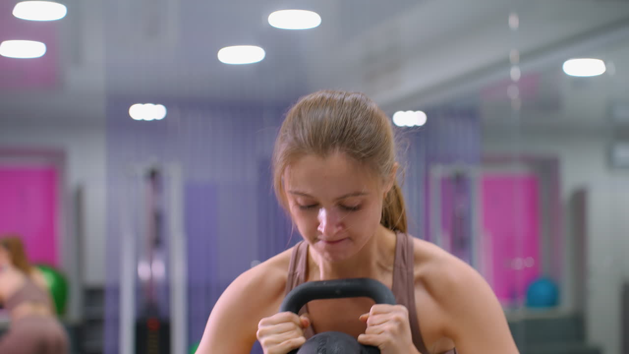 Woman engaging in strength training, lifting kettlebell while stepping onto an elevated surface, then stepping down and repeating, focused workout session in a well-equipped gym environment