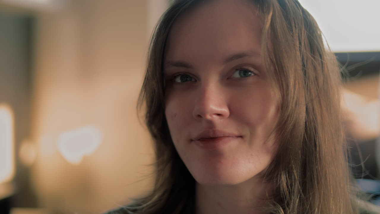 Cinematic close up shot of a beautiful white woman's Face in her apartment looking around and then staring straight into the camera and smiling during a golden hour sunrise in slow motion
