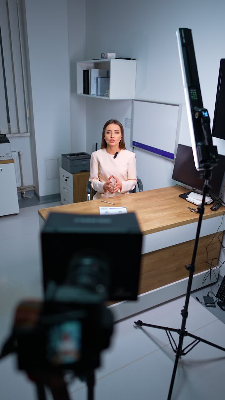 Attractive Caucasian woman sits at desk recording a video for blog. Camera on tripod shooting a modern blogger. High angle view. Vertical video.