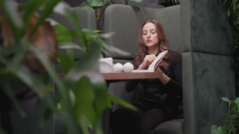 lady in black outfit pulls up sleeve while reaching across table to pink book, surrounded by indoor plants and decoration in peaceful green booth setting with natural tones and soft background focus