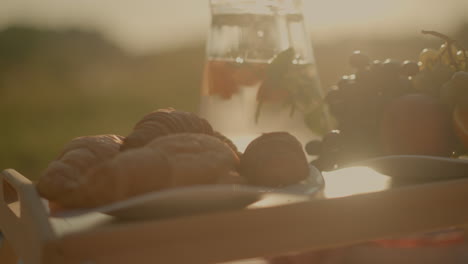 close-up of rustic picnic setup on wooden table featuring fresh croissants, green grapes, and assorted fruits like orange slices and peach, alongside water pitcher under golden