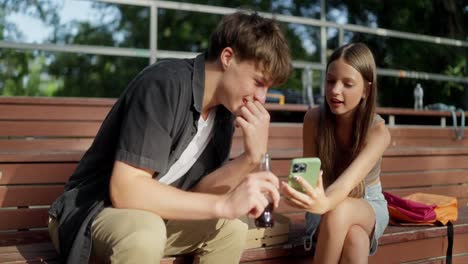 two teenagers sit on a bench, laughing and looking at a phone