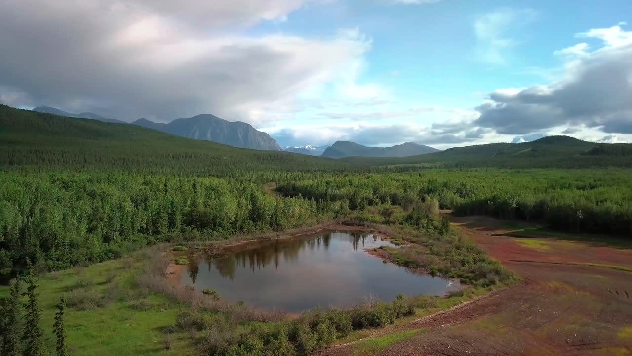 Breathtaking summer Yukon scenic flight above small mirror pond lake by evergreen tree forest Ibex valley towards majestic Mount Ingram, Canada, overhead aerial approach