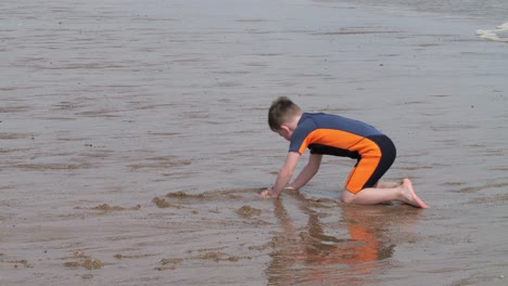 young boy in a wetsuit on a beach digging in the sand