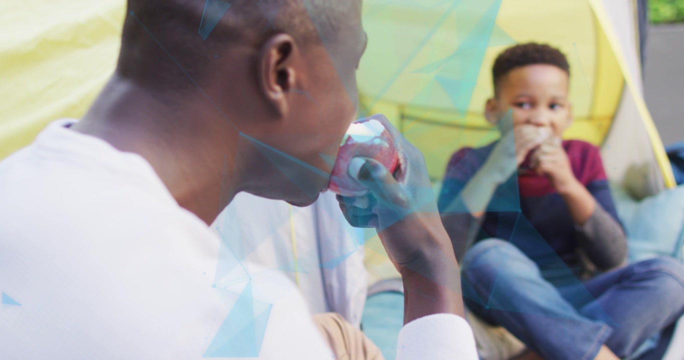 Camera pulling back showing man in white shirt lifting apple, sharing snack beside boy in tent