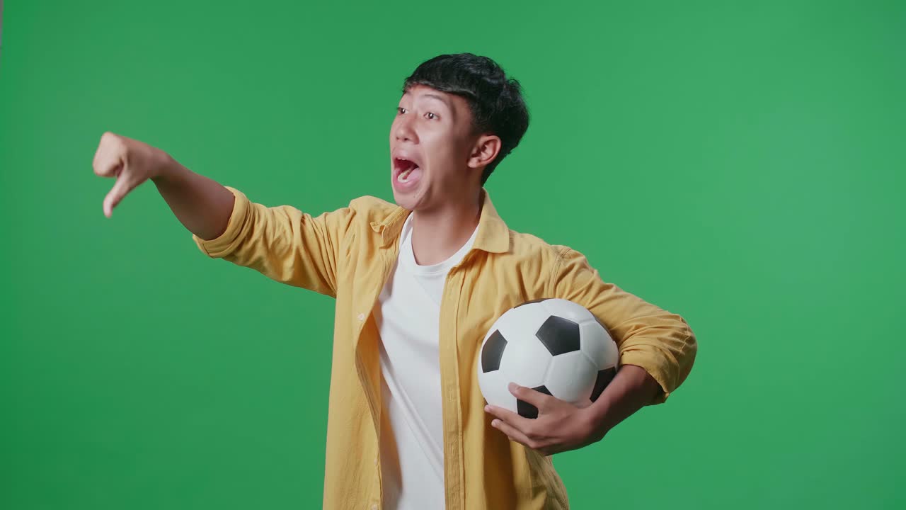 푸른 스크린 배경에서 축구를 응원하는 동안 패자를 조롱하는 몸짓으로 엄지손가락을 아래로 보여주는 공을 가진 아시아 소년의 측면 시각