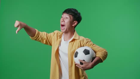 side view of asian boy with a ball showing thumbs down gesture for making fun of the losers while cheering soccer on green screen background