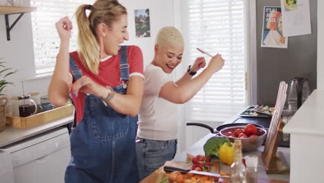 happy diverse female couple cooking vegetables and dancing in kitchen