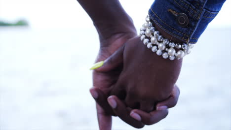 joven pareja afroamericana cogida de las manos por el agua del océano vistiendo joyas en un vestido blanco y chaqueta de vaqueros durante un día nublado cinematográfico 4k