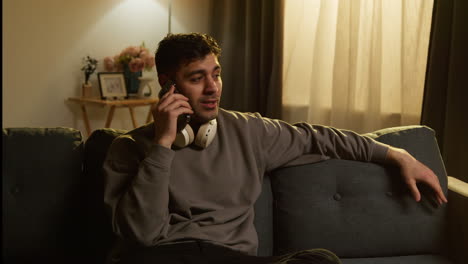 Young-Man-Wearing-Wireless-Headphones-Spending-Evening-At-Home-Sitting-On-Sofa-Talking-On-Mobile-Phone