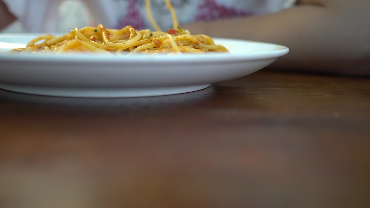 A little girl eats tomato seafood spaghetti in a restaurant. The foreground is blurred and the focus is on the back. The subject is on the left.