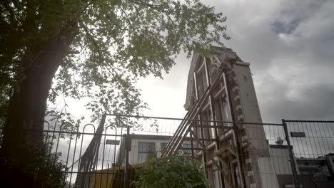 an old fenced off derelict house in amsterdam, with trees swaying in the foreground  and cloudy skies passing over the building.