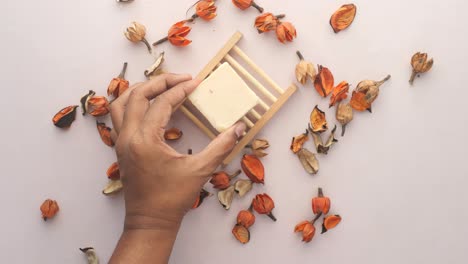 mano recogiendo una barra de jabón en un plato de madera rodeado de flores de naranja secas