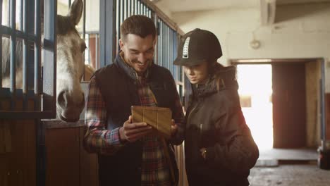 man and young girl are using a tablet computer next to a horse in a stable.