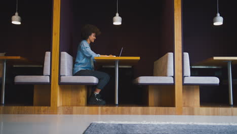 young businesswoman in modern office working on laptop in seating pod with takeaway drink