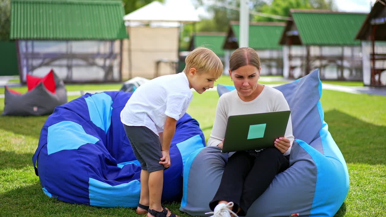 Woman showing something weird on laptop to a blond boy beside her. Kid comes up, looks with interest and comments.