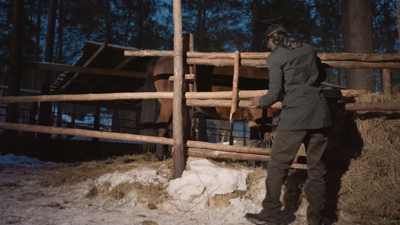 reloj de madera nocturno, guardia nocturno revisa recinto nevado, inspección crepuscular del recinto rústico de madera a la luz de la linterna, cuidador observa el cercado cubierto de nieve al anochecer con una postura concentrada
