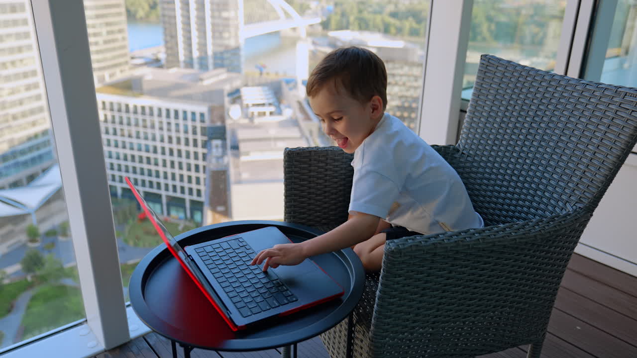 Little boy working on red laptop on balcony. Small boy concentrating while using laptop on high-rise balcony with cityscape view